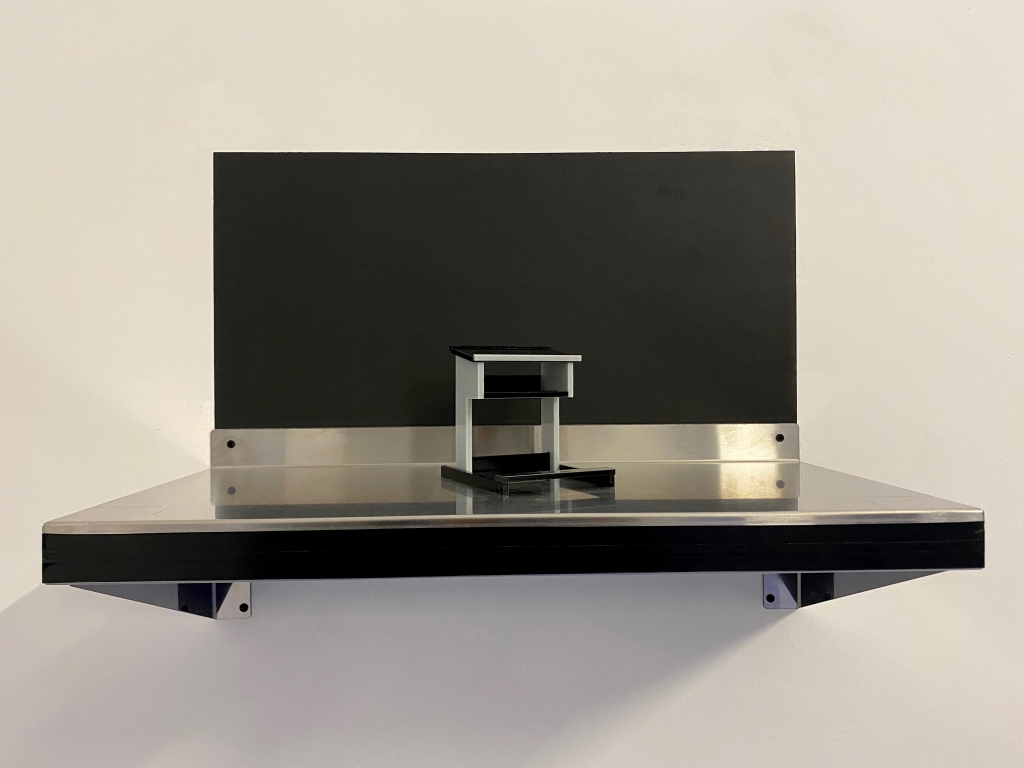 A model, grey and black plastic sits on a metal shelf. A square of dark paint is behind the model and shelf.